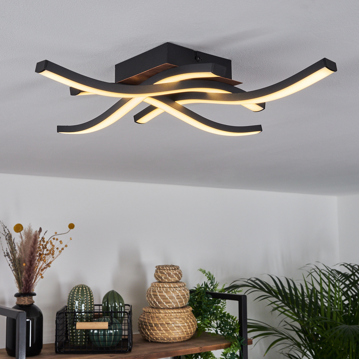 Eine moderne Deckenlampe mit geschwungenen schwarzen Linien und warmweißen Lichtern. Regale mit Deko.