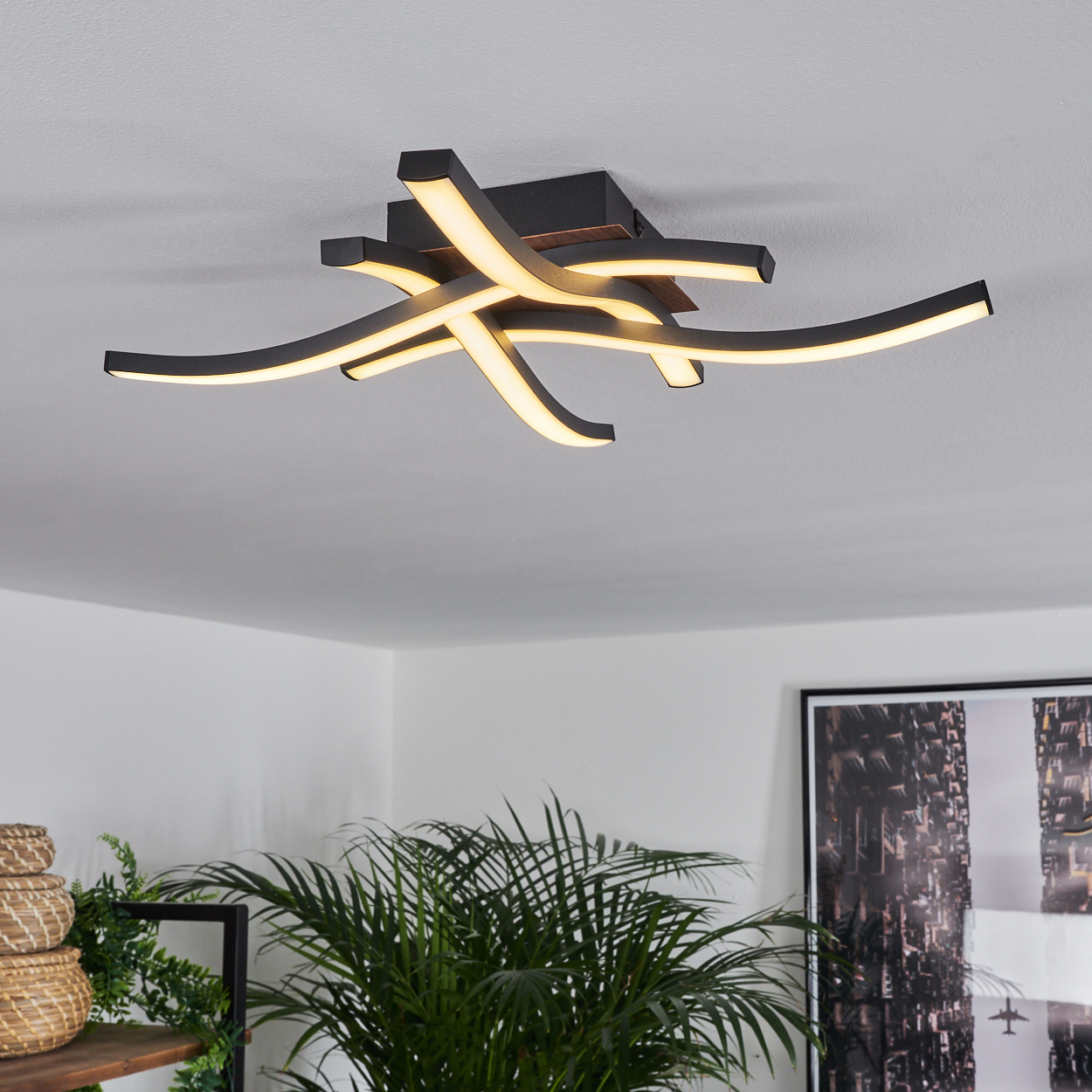 Moderne Deckenleuchte mit gebogenen, beleuchteten Armen. Dunkle Decke, weiße Wand und Pflanze im Hintergrund.