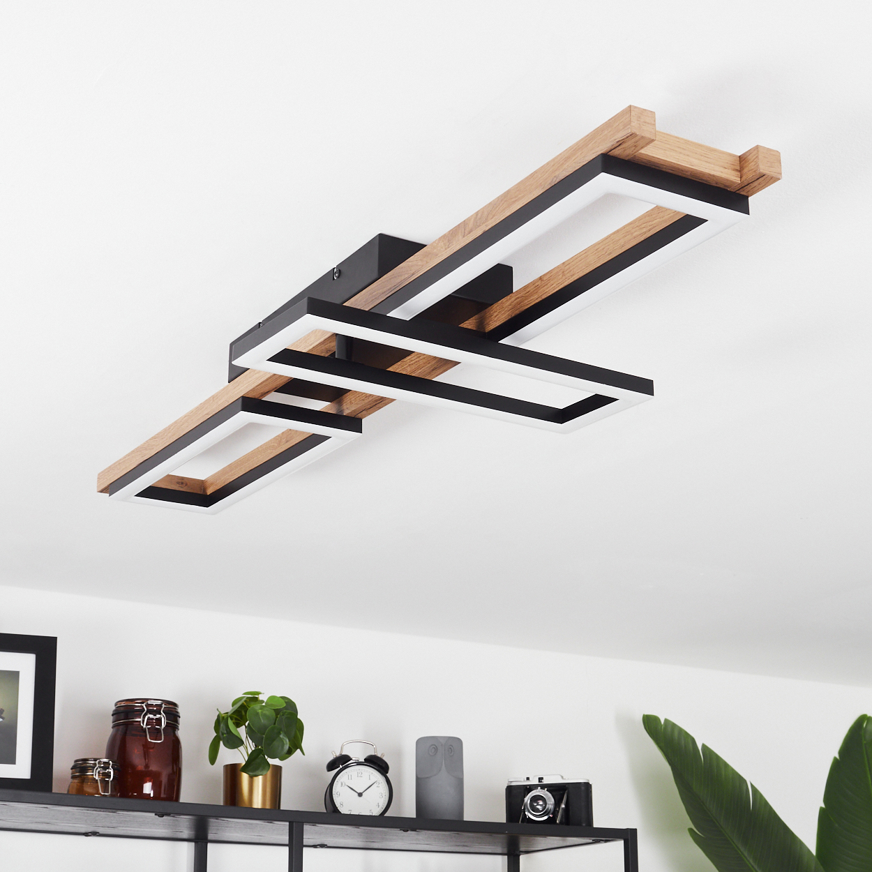 Deckenleuchte mit Holz- und schwarzen Rechteckrahmen mit weißen Lichtstreifen. Darunter ein Regal mit Dekorationen.
