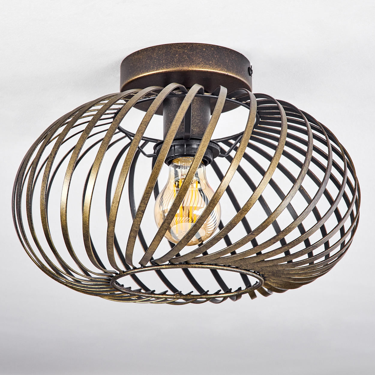 Deckenlampe mit spiralförmigem Drahtrahmen. Eine Glühbirne ist sichtbar. Der Sockel ist bronze. Weiße Decke.