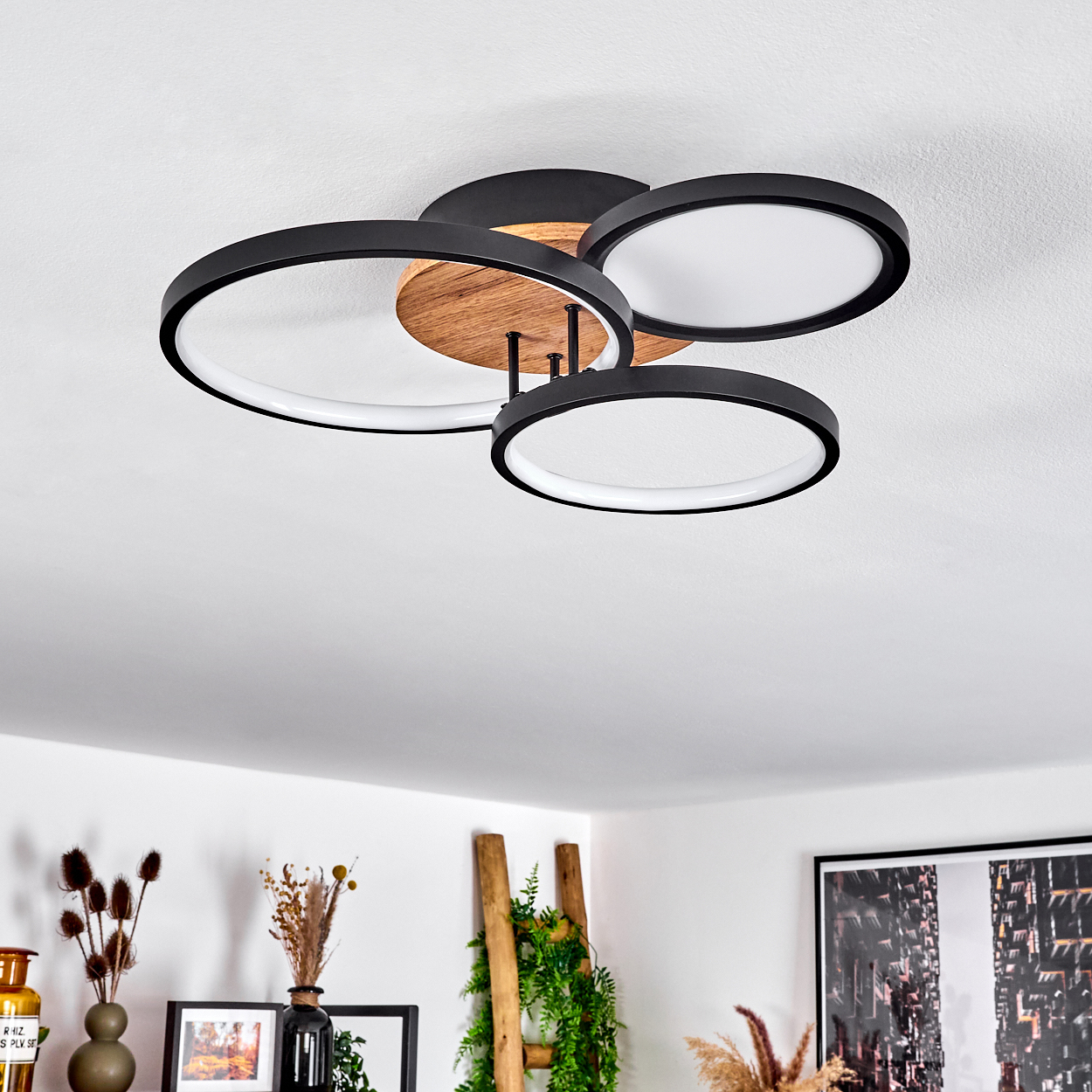 Deckenlampe mit drei schwarzen, ovalen Leuchten und Holzakzenten. Der Hintergrund zeigt eine Leiter und Dekoration.