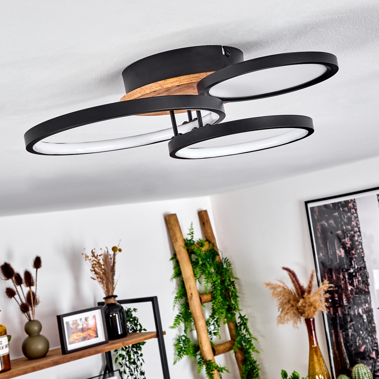 Moderne Deckenlampe mit drei schwarzen, ovalen Leuchten und Holzakzenten. Der Hintergrund zeigt Dekoration und eine Leiter.