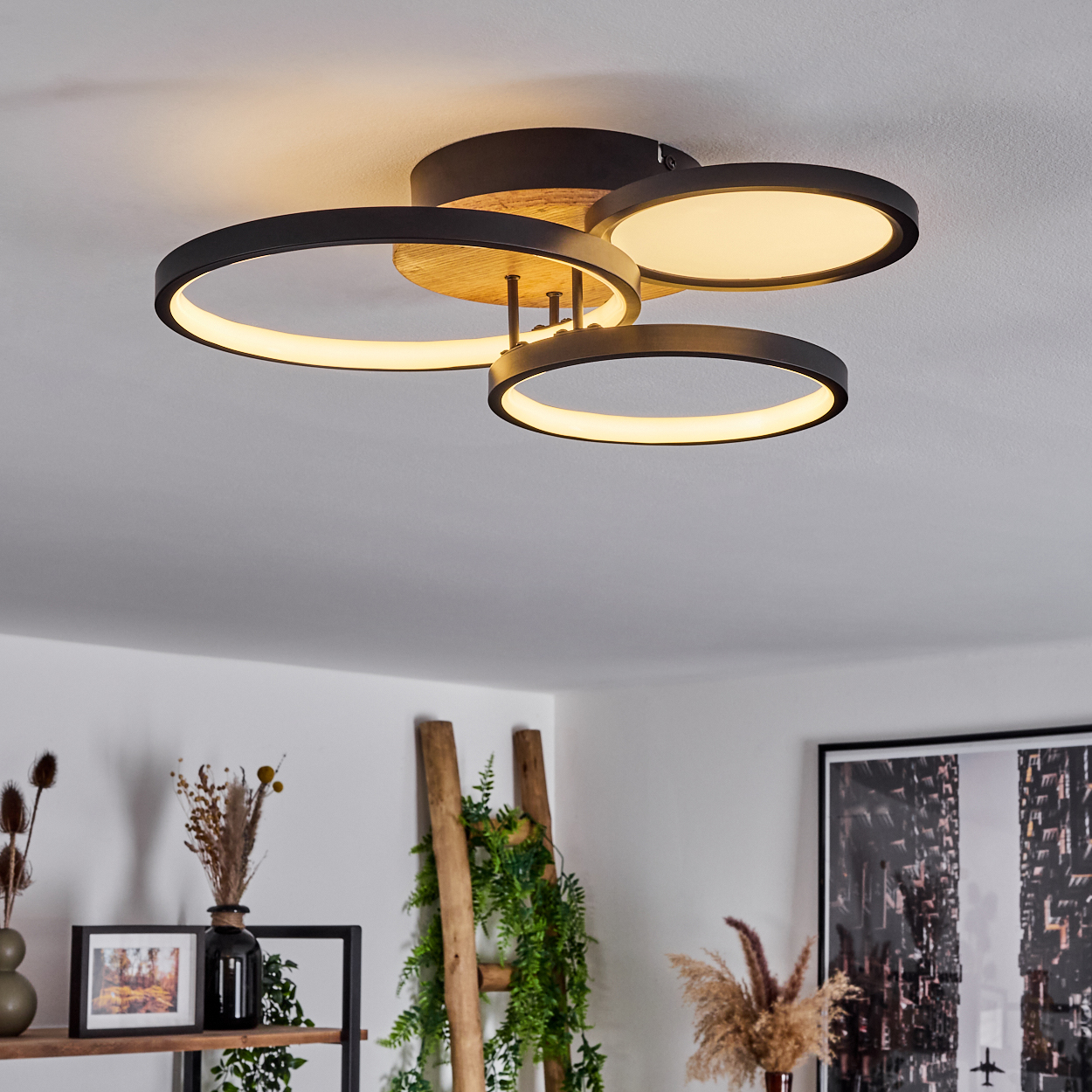 Deckenlampe mit drei schwarzen, ovalen Leuchten und Holzakzenten. Der Hintergrund zeigt Dekoration.