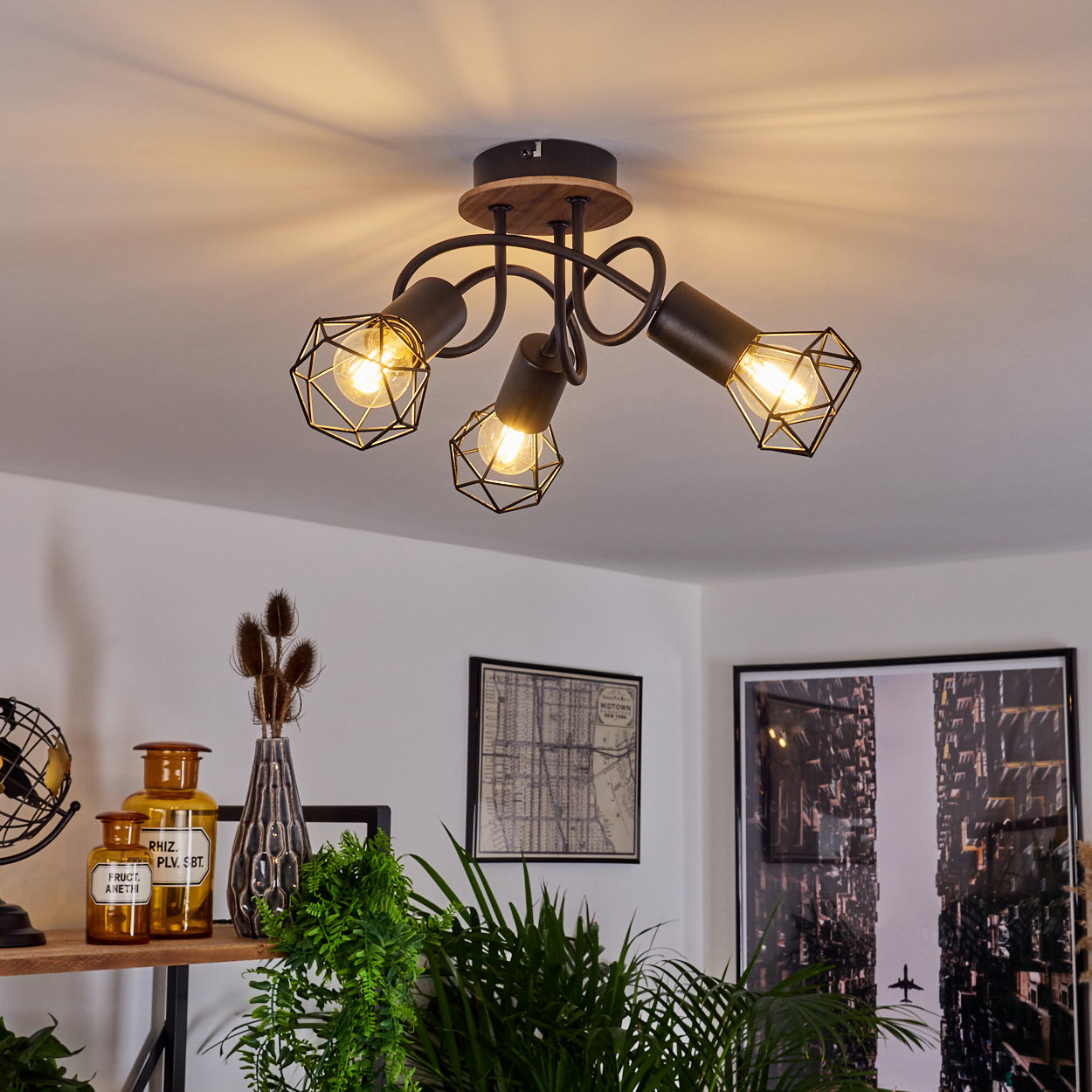 Eine Deckenleuchte mit drei abgewinkelten Lampen, in einem Raum mit Pflanzen und Bildern an der Wand.