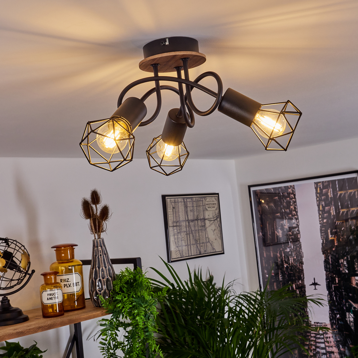 Eine Deckenleuchte mit drei abgewinkelten Lampen, in einem Raum mit Pflanzen und Bildern an der Wand.