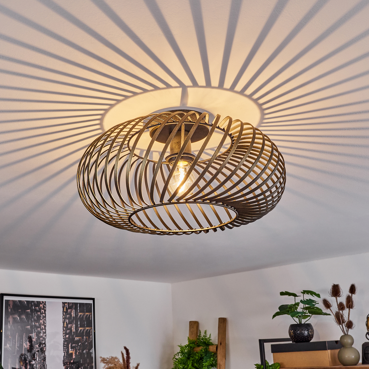 Eine bronzefarbene, spiralförmige Deckenlampe hängt, ihr Schatten wird auf die Decke geworfen. Innenelemente wie Pflanzen und Kunst sind sichtbar.