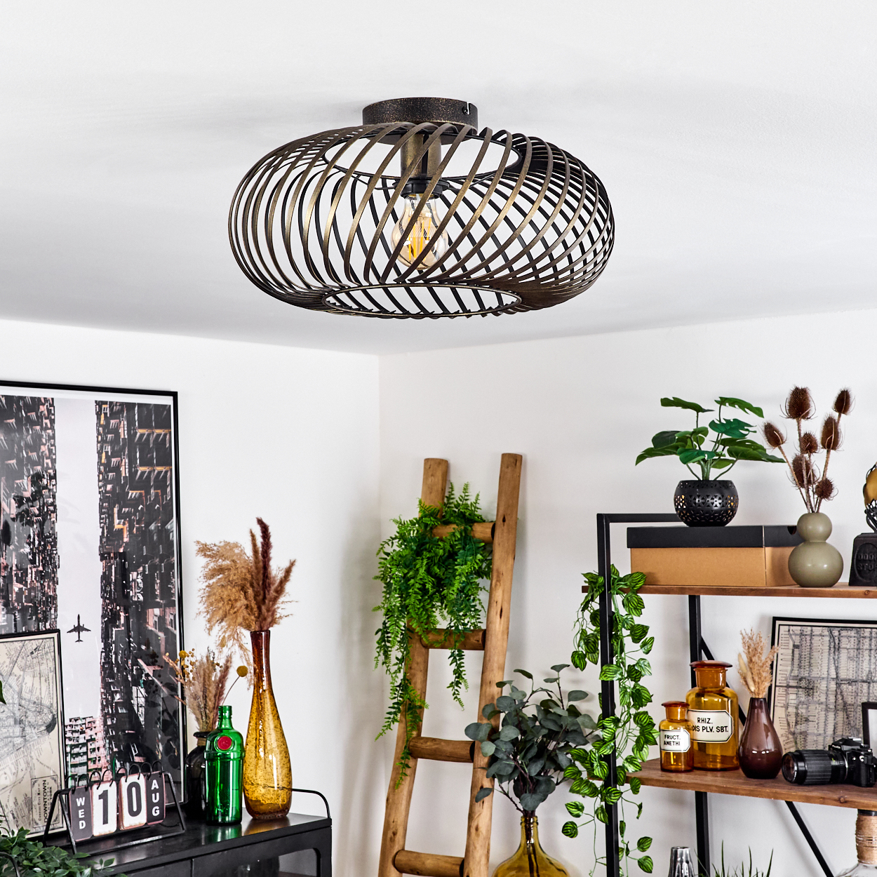 Eine bronzefarbene Spiral-Deckenlampe hängt. Der Innenraum umfasst Pflanzen, eine Leiter und Kunstwerke an einer weißen Wand.