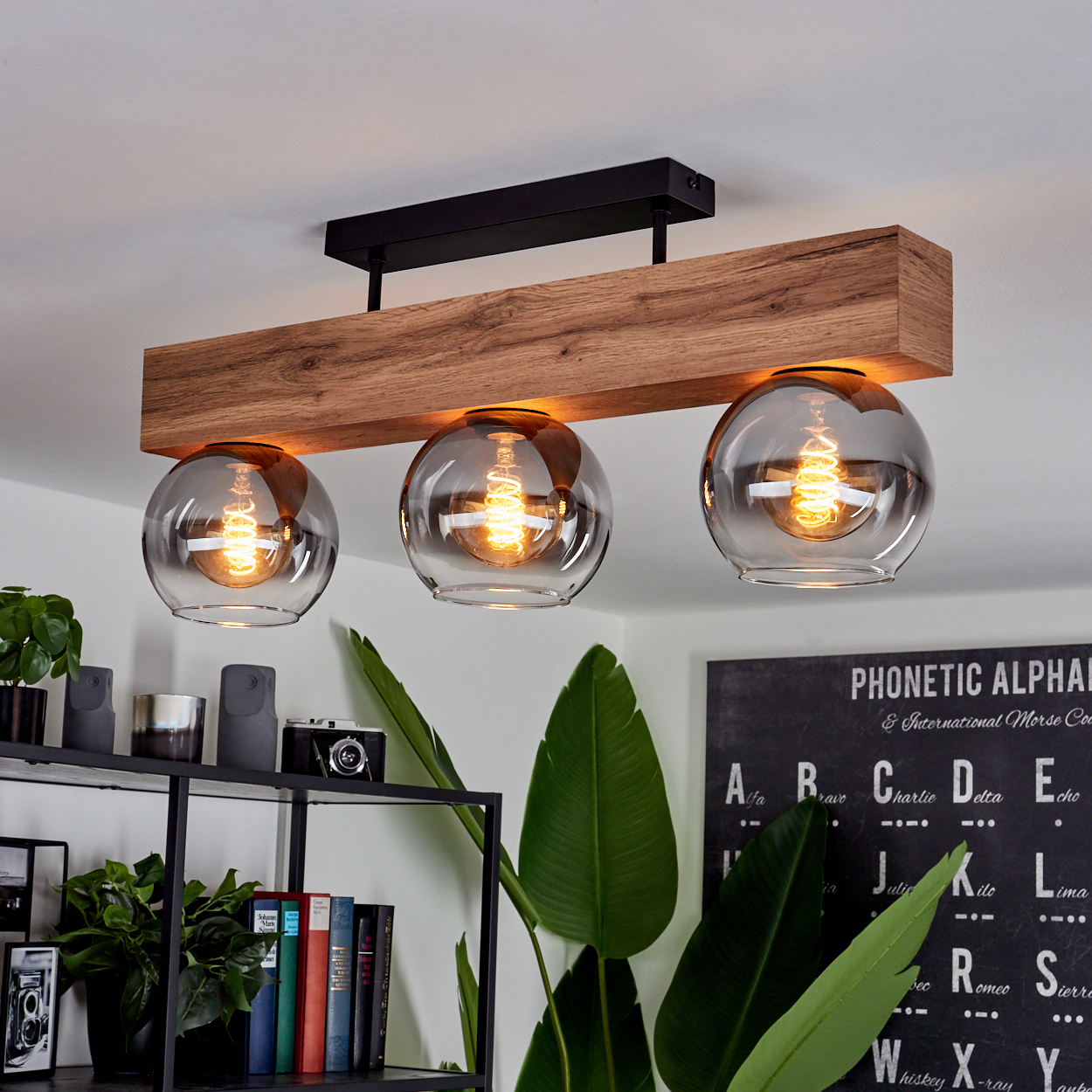 Ein Holzbalken mit drei Glaskugellichtern, die Lichter sind an. Eine Tafel und Pflanzen im Hintergrund.