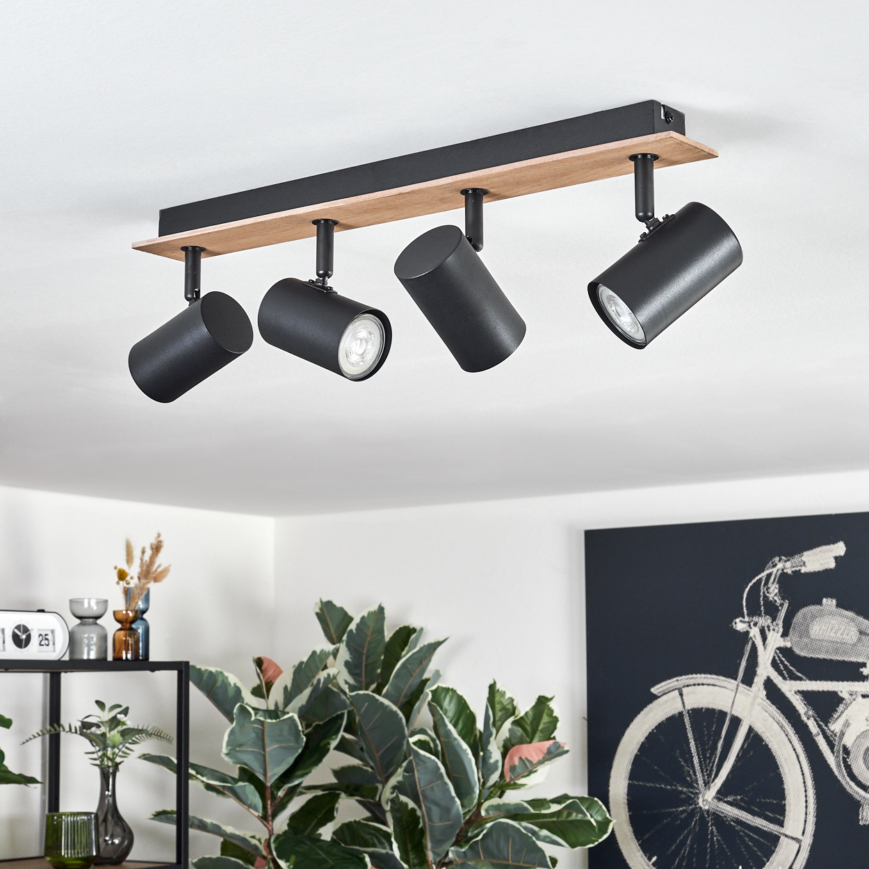 Vier schwarze Scheinwerfer hängen von einer hellen Holzbasis an einer weißen Decke in einem Raum.