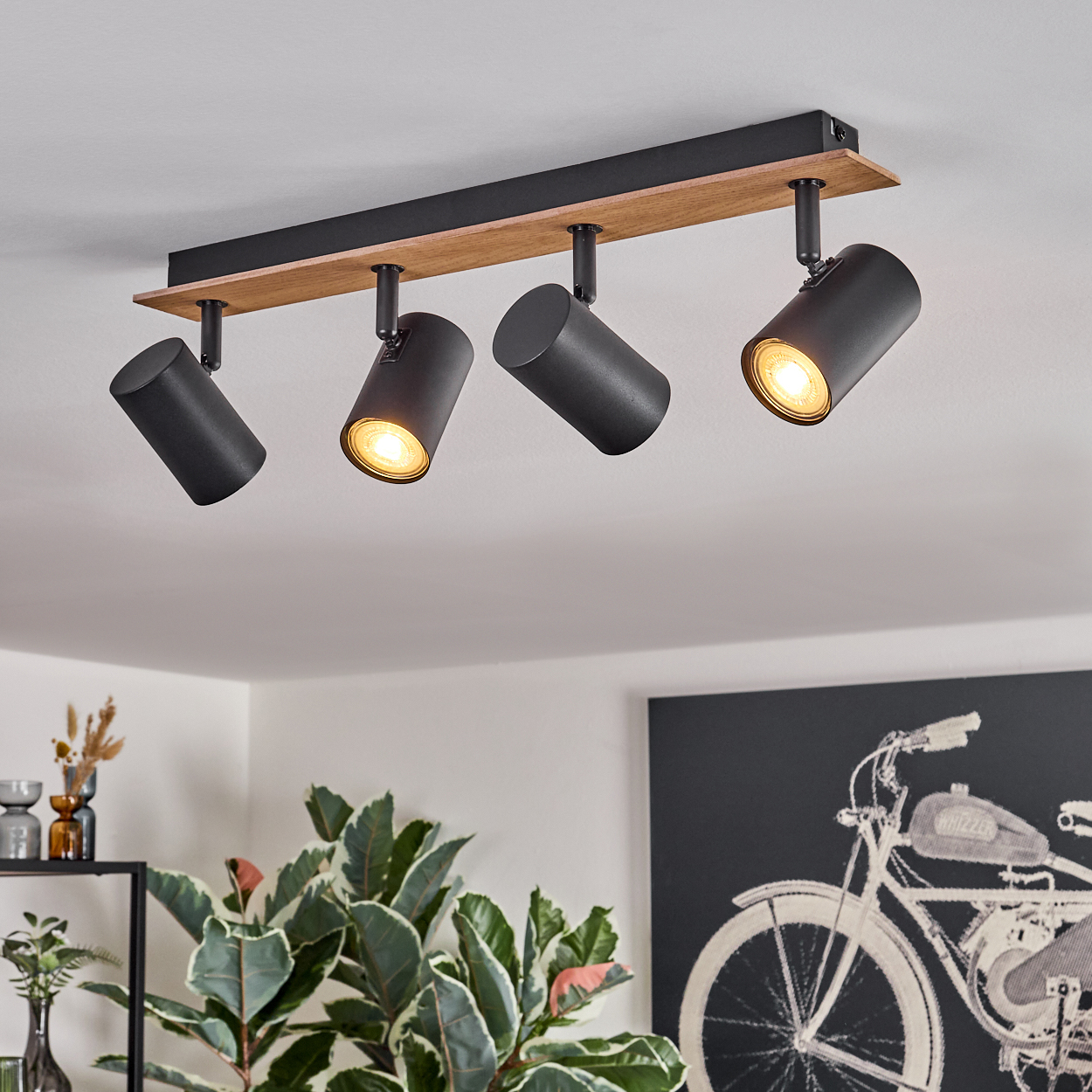 Vier schwarze Scheinwerfer hängen von einer hellen Holzbasis an einer weißen Decke in einem Raum.