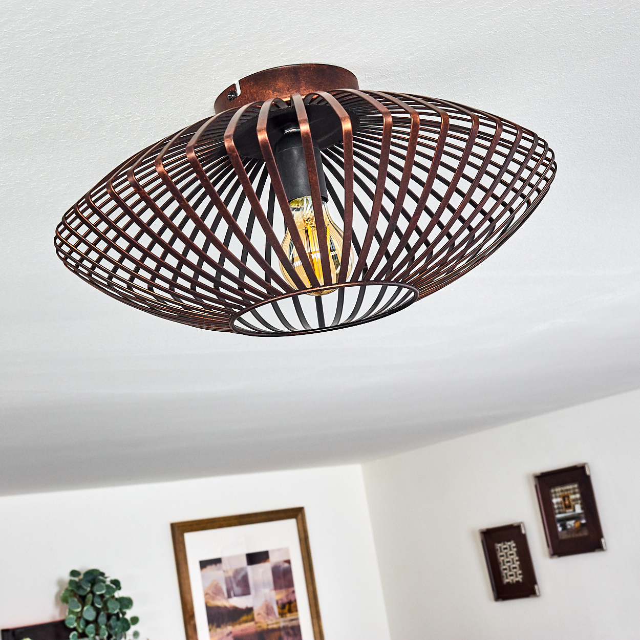 Bronzefarbene Käfig-Deckenleuchte mit sichtbarer Glühbirne, vor weißer Decke.