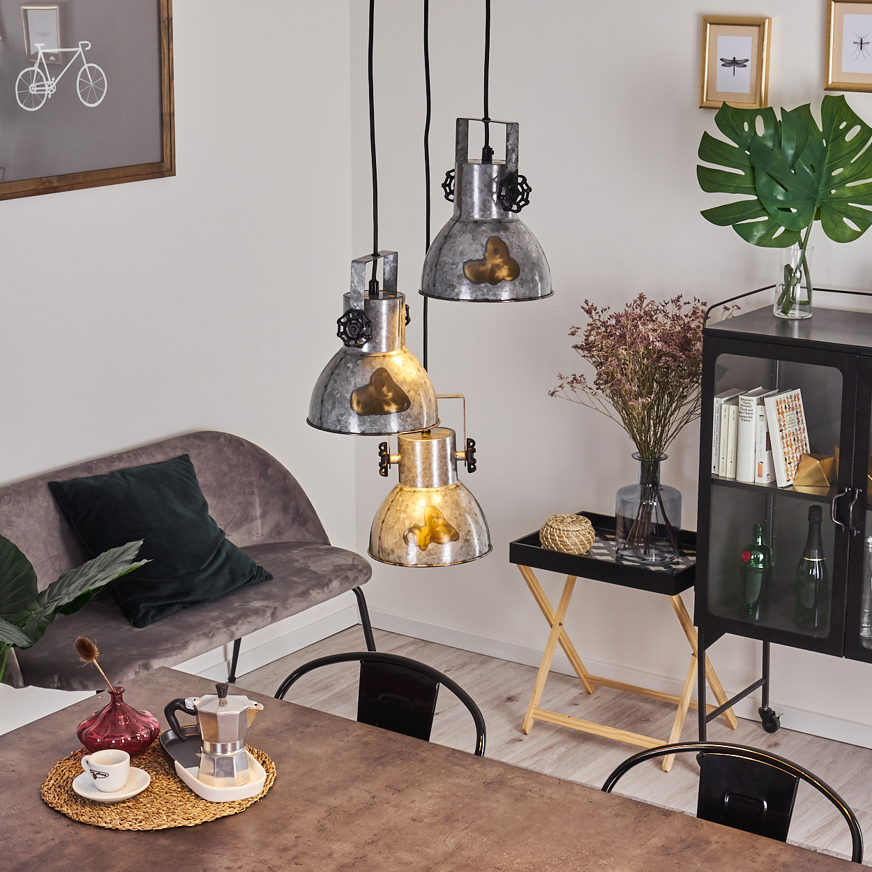 Ein Esszimmer mit drei Metallpendelleuchten über einem Tisch mit einer Kaffeemaschine und grauen Sitzgelegenheiten.