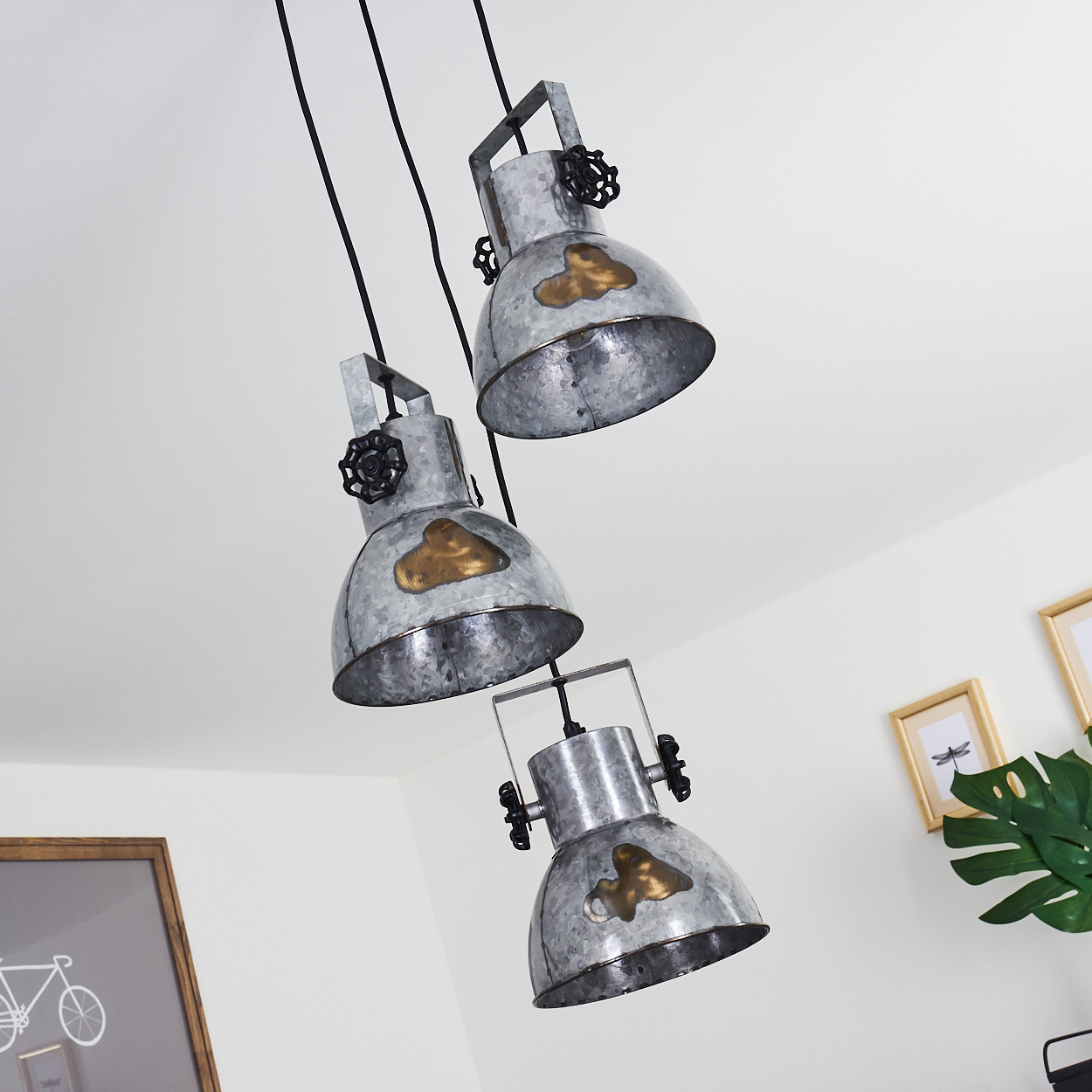 Drei Metallpendelleuchten hängen von einer weißen Decke und beleuchten einen Raum mit gerahmter Kunst.