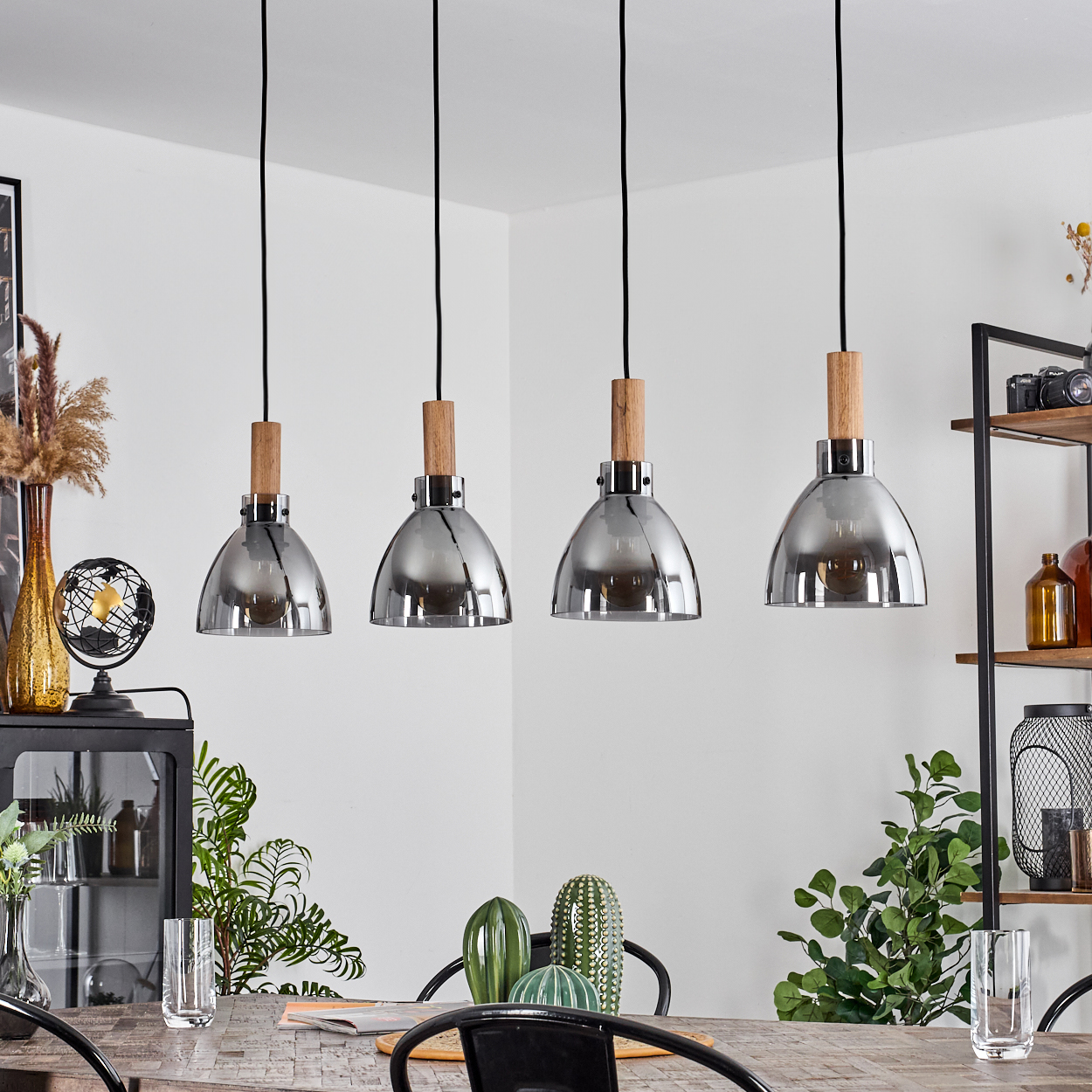 Vier Pendelleuchten. Sie hängen über einem Tisch mit Pflanzen und Gläsern. Die Leuchten sind chromfarben mit Holz.