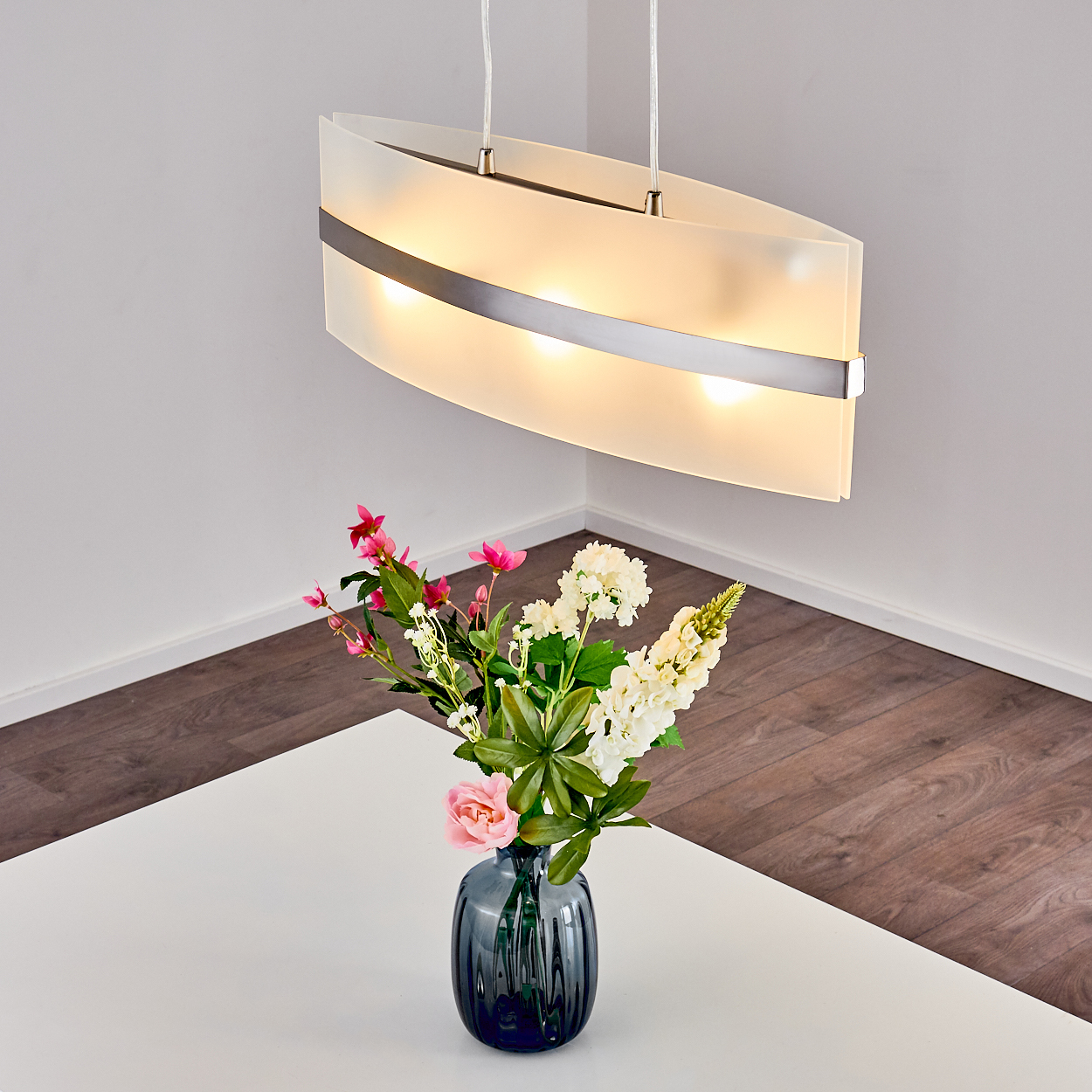 Weiße Deckenlampe über einem Tisch mit Blumen in einer dunklen Vase. Die Lampe ist eingeschaltet.