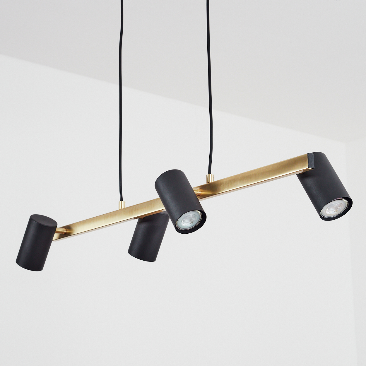 Eine Leuchte mit einem goldenen Balken und vier schwarzen Zylinderleuchten, abgewinkelt und beleuchtet, vor weißem Hintergrund.