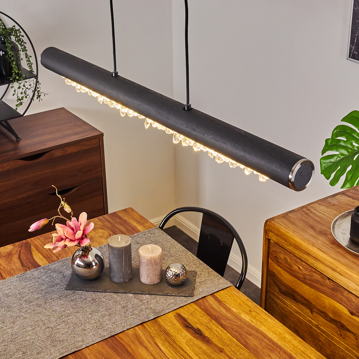 Eine Esszene. Über einem Tisch hängt eine lange, schwarze Lampe mit Kristalltropfen. Kerzen und Dekor sind vorhanden.