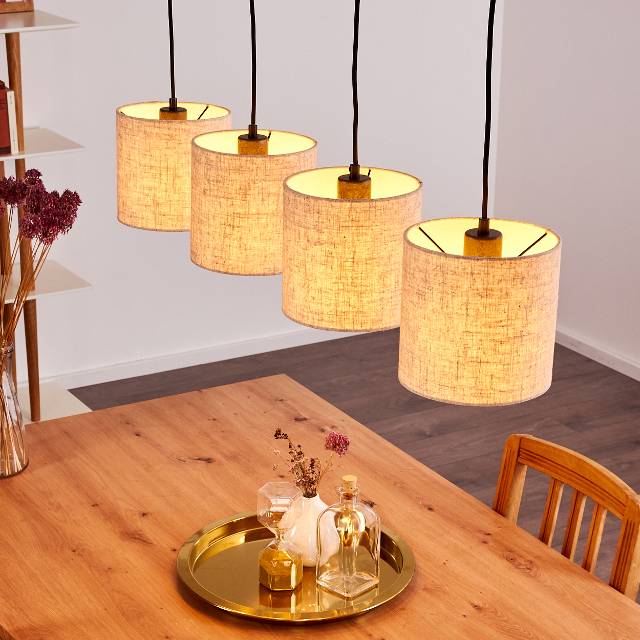 Vier Pendelleuchten mit zylindrischen, beigen Schirmen hängen über einem Holztisch. Die Leuchten sind beleuchtet.