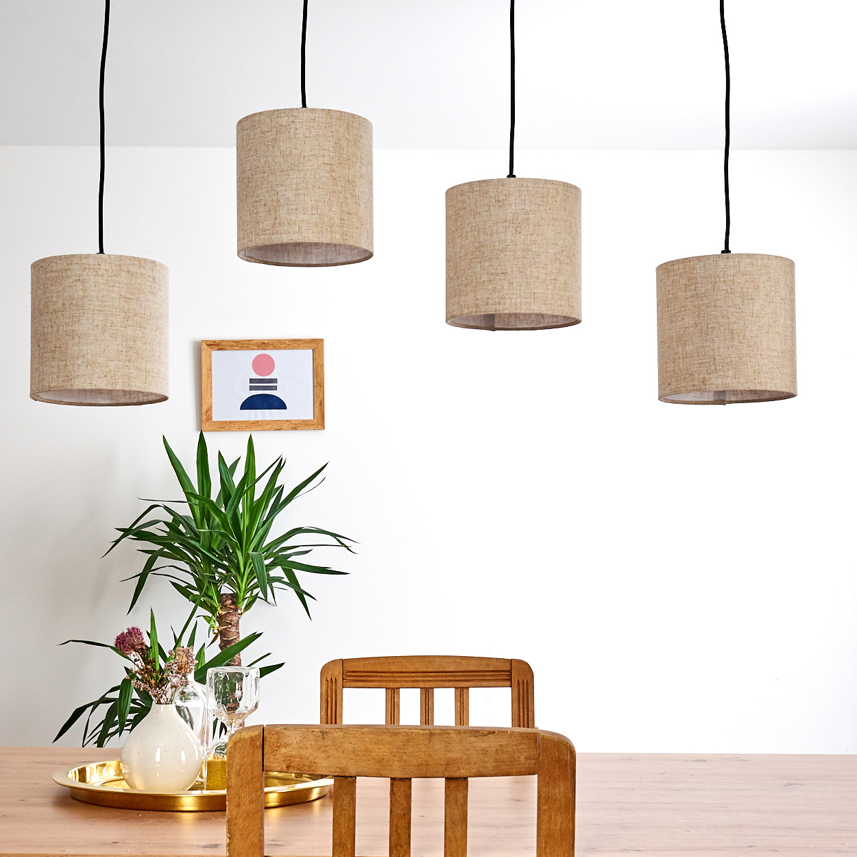 Vier Pendelleuchten mit beigen Schirmen hängen über einem Holztisch. Ein Gemälde, eine Vase und ein Stuhl sind in der Szene.