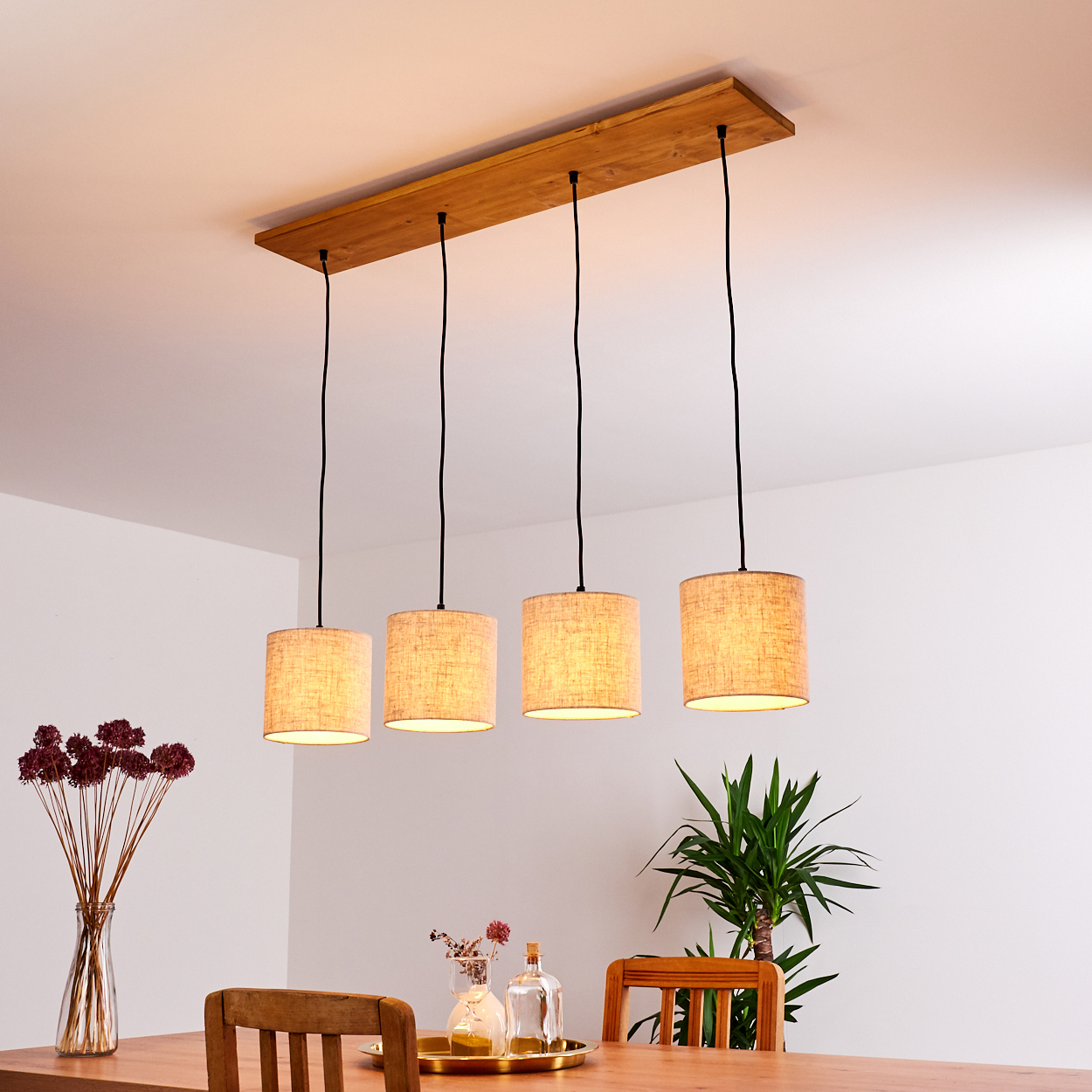 Vier Pendelleuchten mit beigen Schirmen hängen über einem Holztisch. Eine Vase und Blumen sind ebenfalls vorhanden.