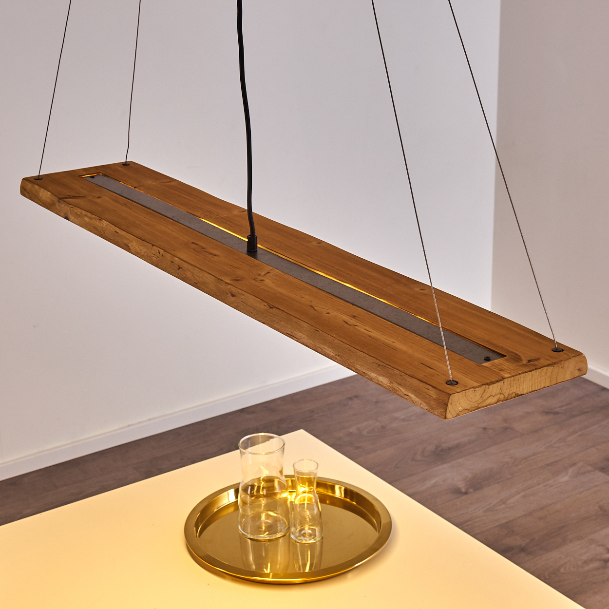 Eine Holzbalken-Deckenleuchte mit zentralem Lichtstreifen, hängt über einem Tisch.