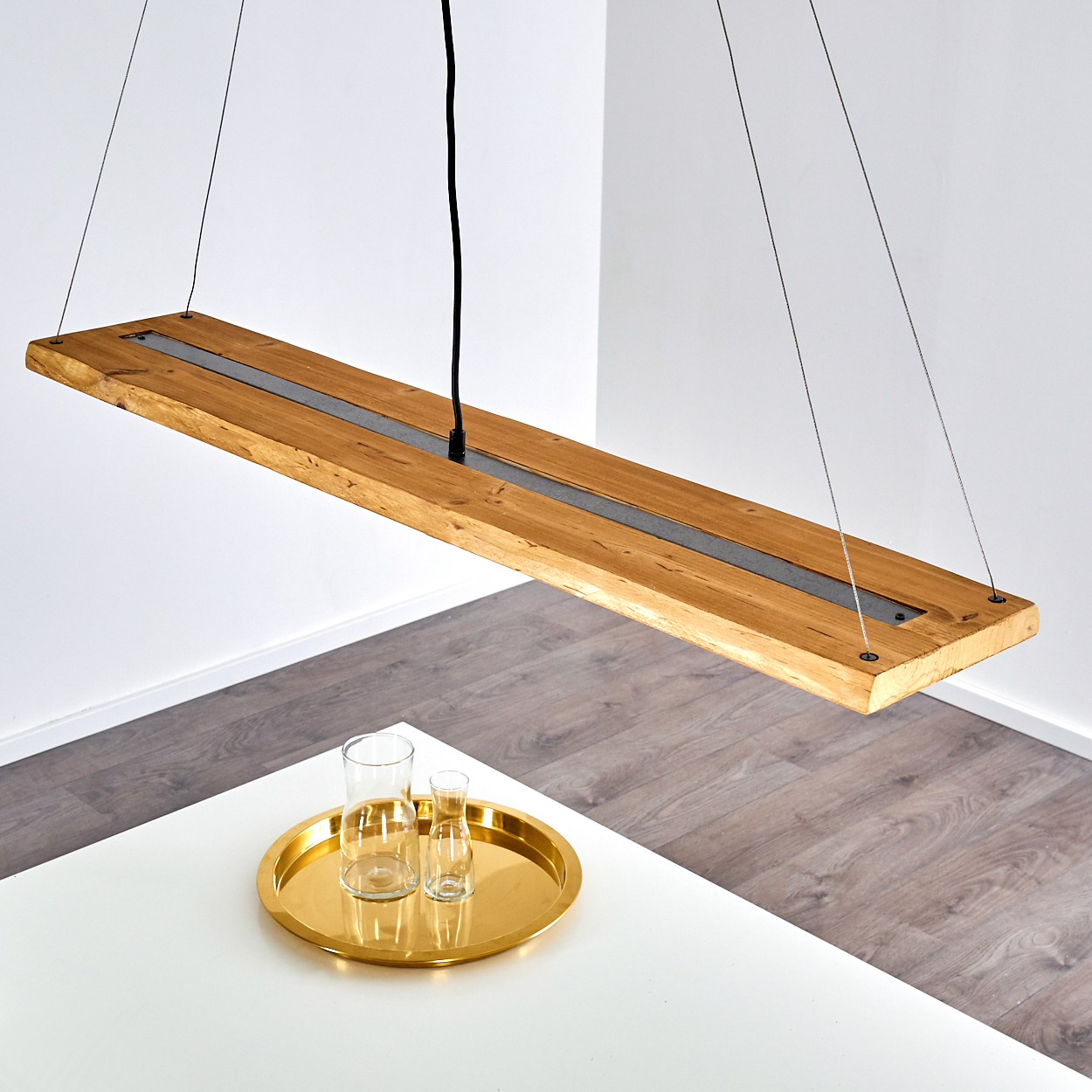 Hölzerne, rechteckige Pendelleuchte mit einem zentralen dunklen Streifen, die über einem weißen Tisch mit goldenem Tablett und Gläsern hängt.