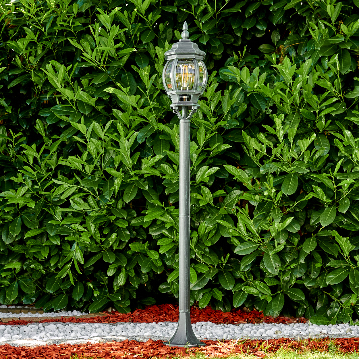 Gartenlaternenpfahl mit klarer Glaskugel und dekorativer Spitze vor einer grünen Hecke.