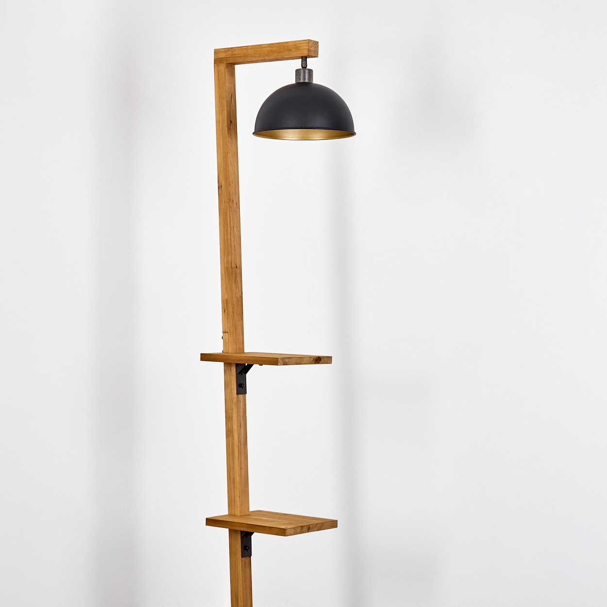 Eine hohe hölzerne Stehlampe verfügt über einen schwarzen Lampenschirm mit goldener Innenseite und zwei kleine Regale am Holzrahmen.