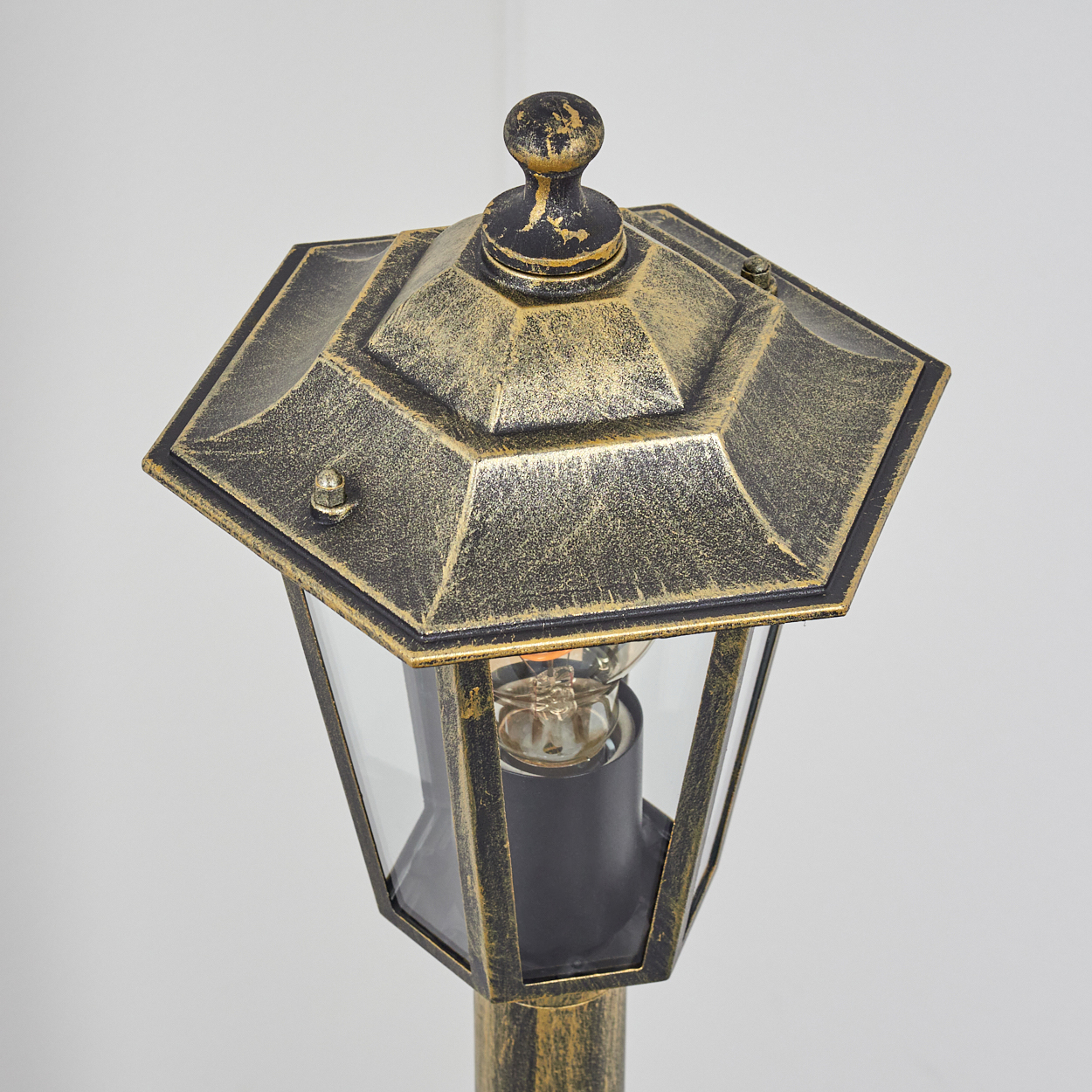 Eine Nahaufnahme einer bronzefarbenen Außenlampe mit sechseckigem Oberteil und klaren Glasscheiben, die die Glühbirne im Inneren zeigt.
