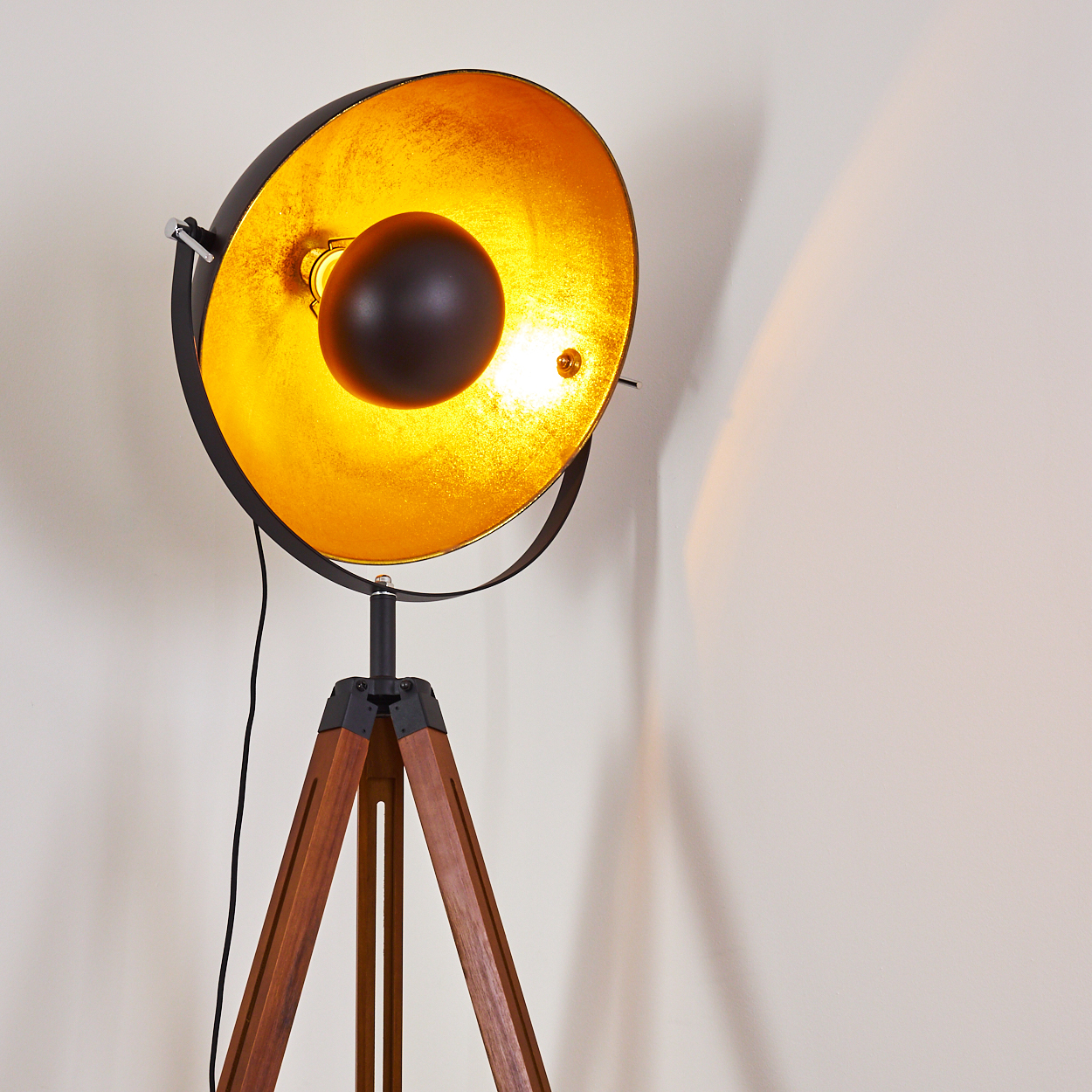 Stativ-Stehleuchte mit schwarz-goldenem Schirm. Holzbeine. Licht ist an.