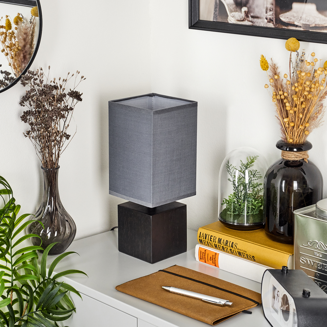 Quadratische Lampe mit schwarzem Sockel und grauem Schirm, neben Spiegel und Dekoration.