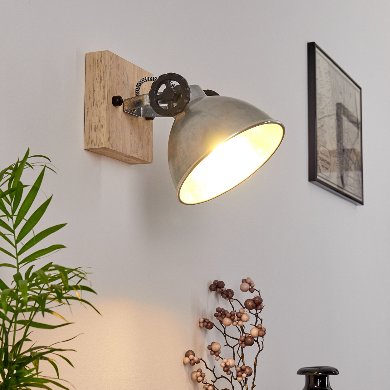 Eine metallische Wandleuchte an einem Holzblock montiert. Das Licht ist an und scheint hell. Pflanze und Bild sichtbar.