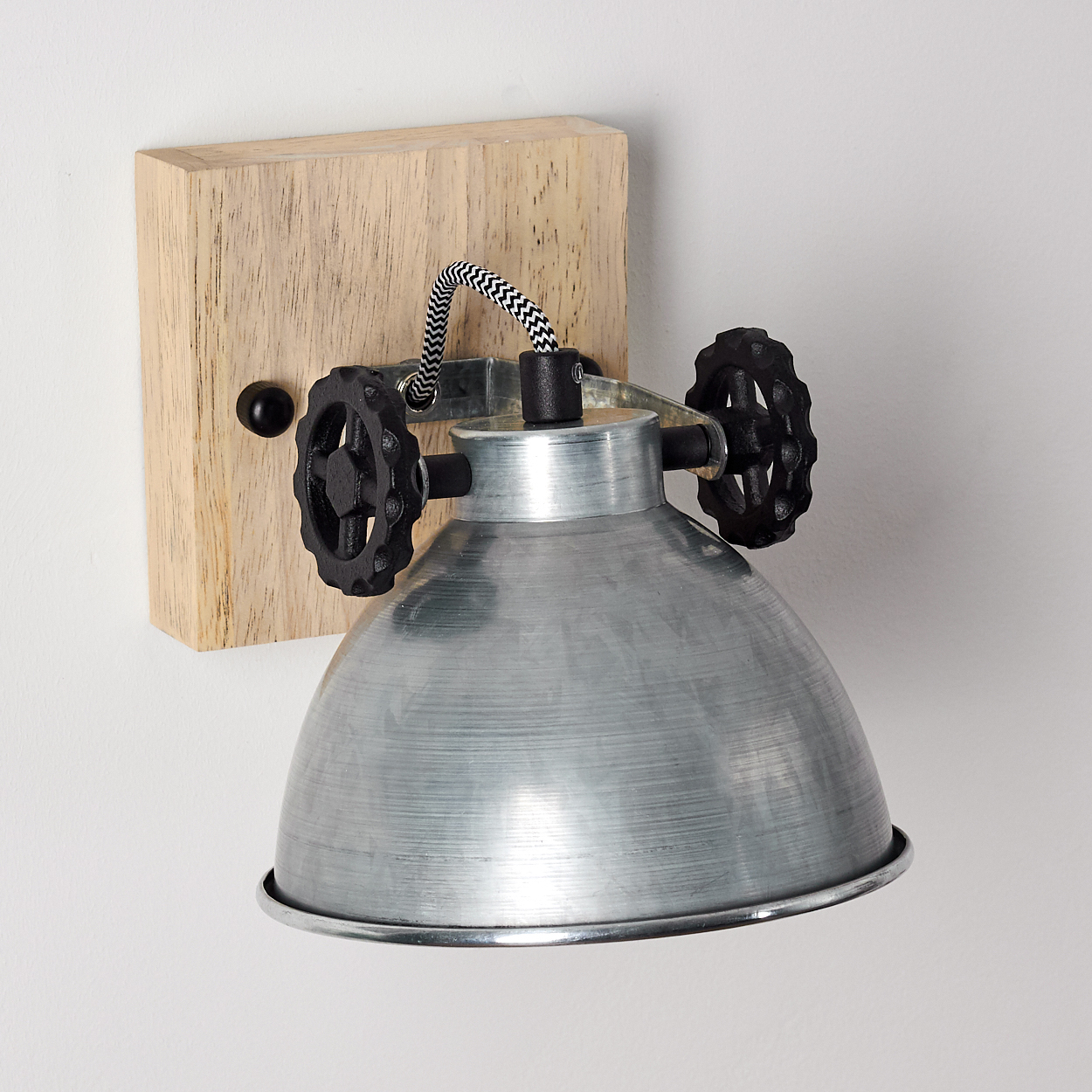 Silberne Metallwandlampe mit schwarzen Zahnradakzenten, montiert auf einem hellen Holzsockel vor einer weißen Wand.