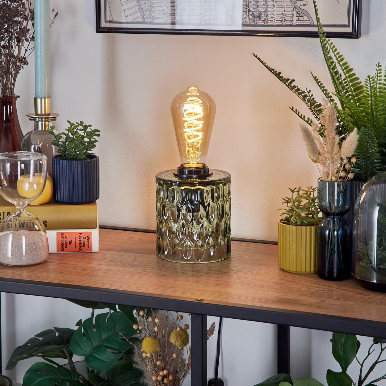Eine dekorative Tischlampe mit einem gemusterten Glassockel, einer Glühbirne im Vintage-Stil und umgeben von Pflanzen und Büchern.