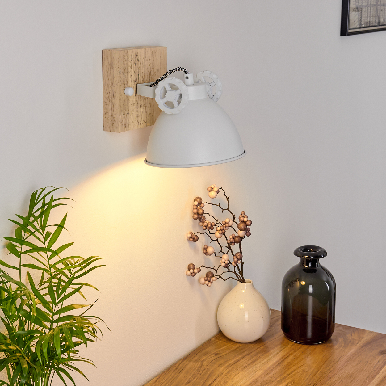 Weiße Wandlampe mit Holzsockel, beleuchtet, nach unten strahlend. Blumen und Vase sind sichtbar.