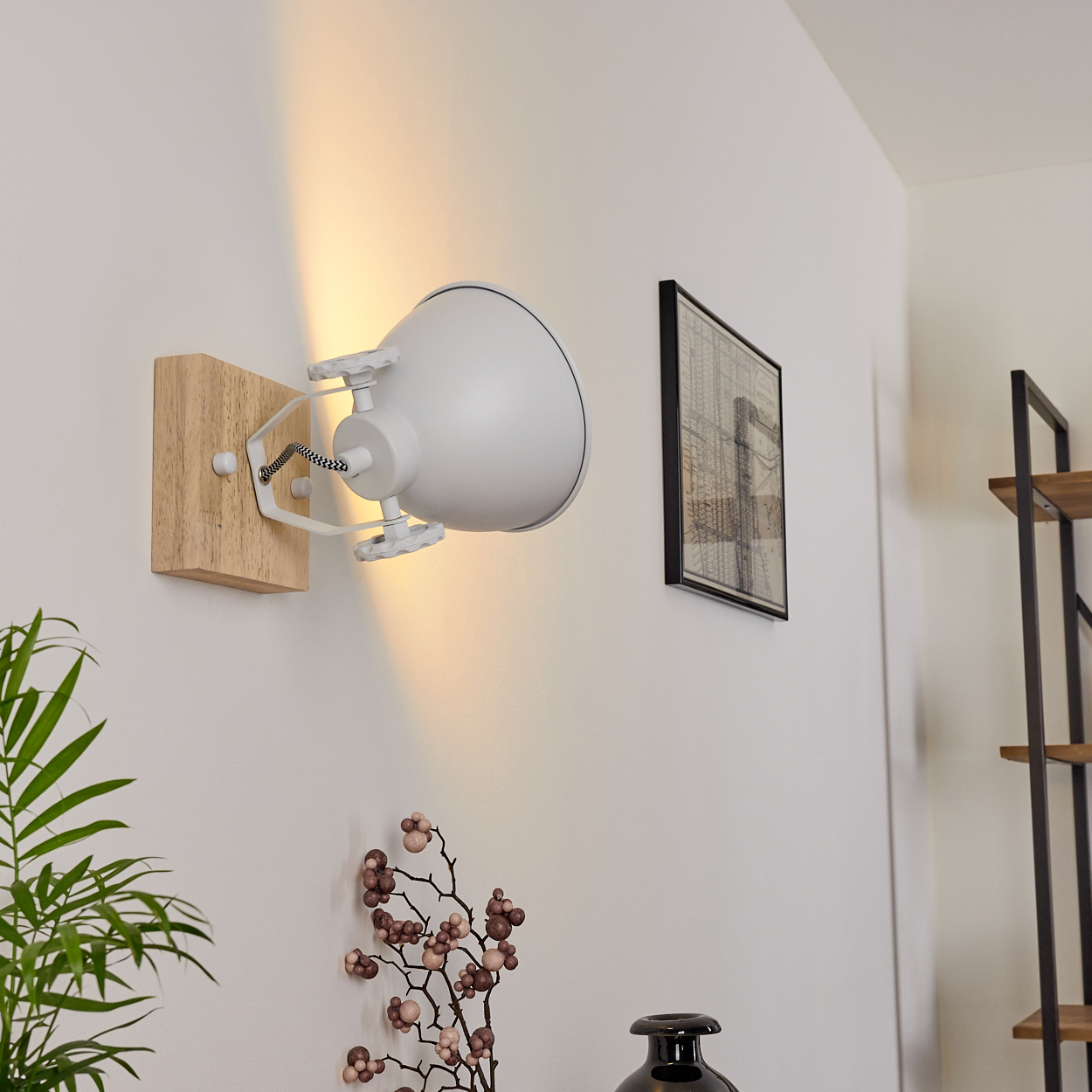 Weiße Wandlampe mit Holzsockel. Die Lampe leuchtet nach unten. Eine Pflanze und ein Bild sind sichtbar.