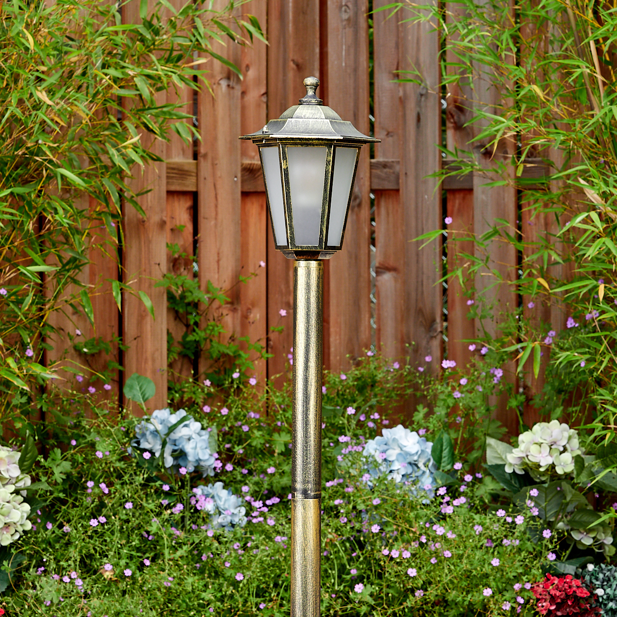 Eine goldene Außenlampe steht in einem Garten. Dahinter ein Holzzaun und grüne Pflanzen. Blumen sind im Vordergrund.