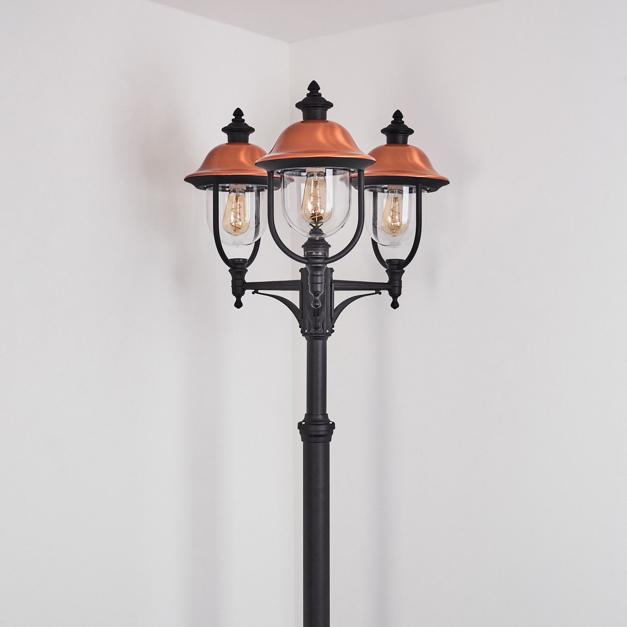 Eine Laterne mit drei Lichtern, mit Kupferaufsätzen, vor einer weißen Wand in einer Ecke.