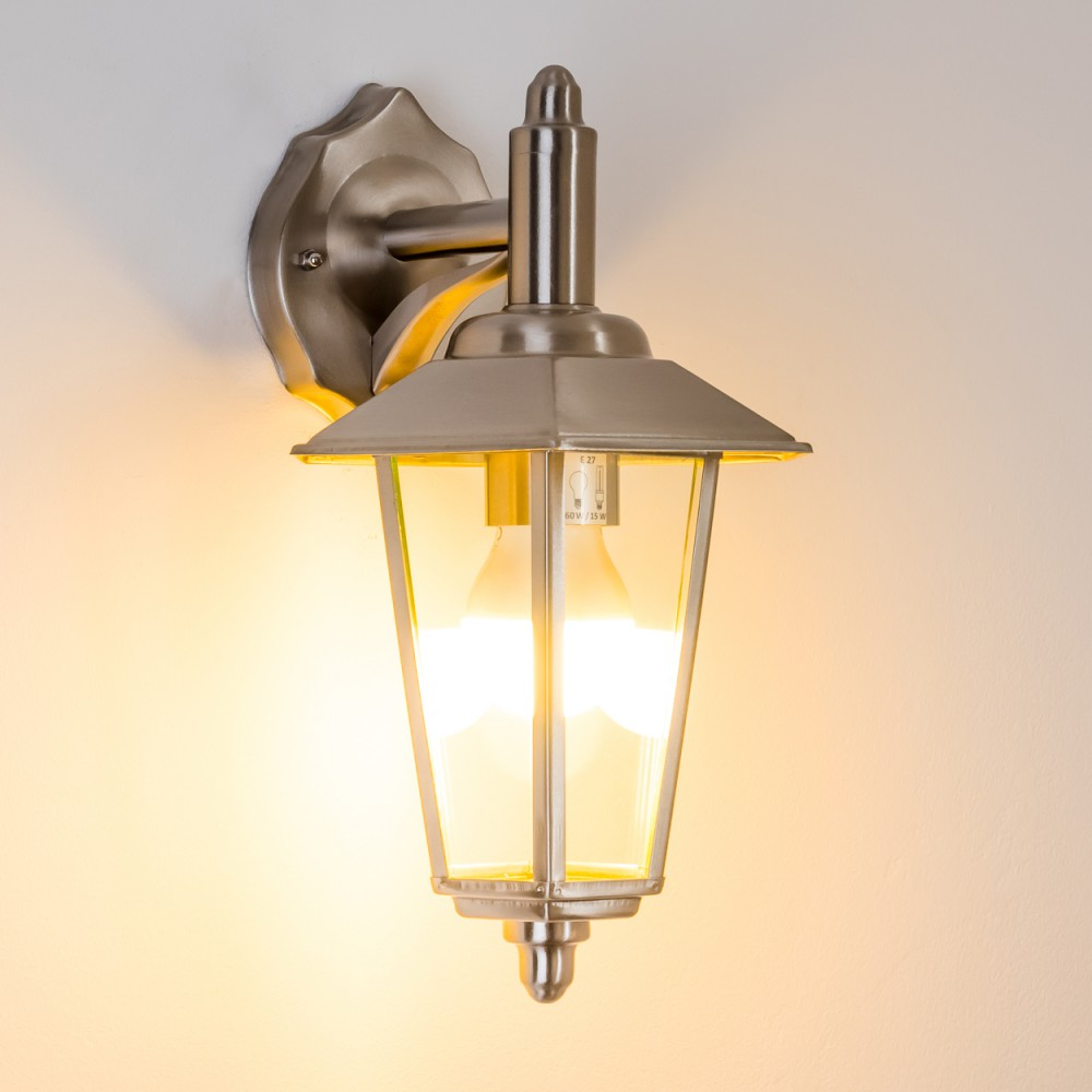Beleuchtete silberne Außenwandleuchte mit klarem Glas, die ein warmes Licht auf eine weiße Wand wirft.