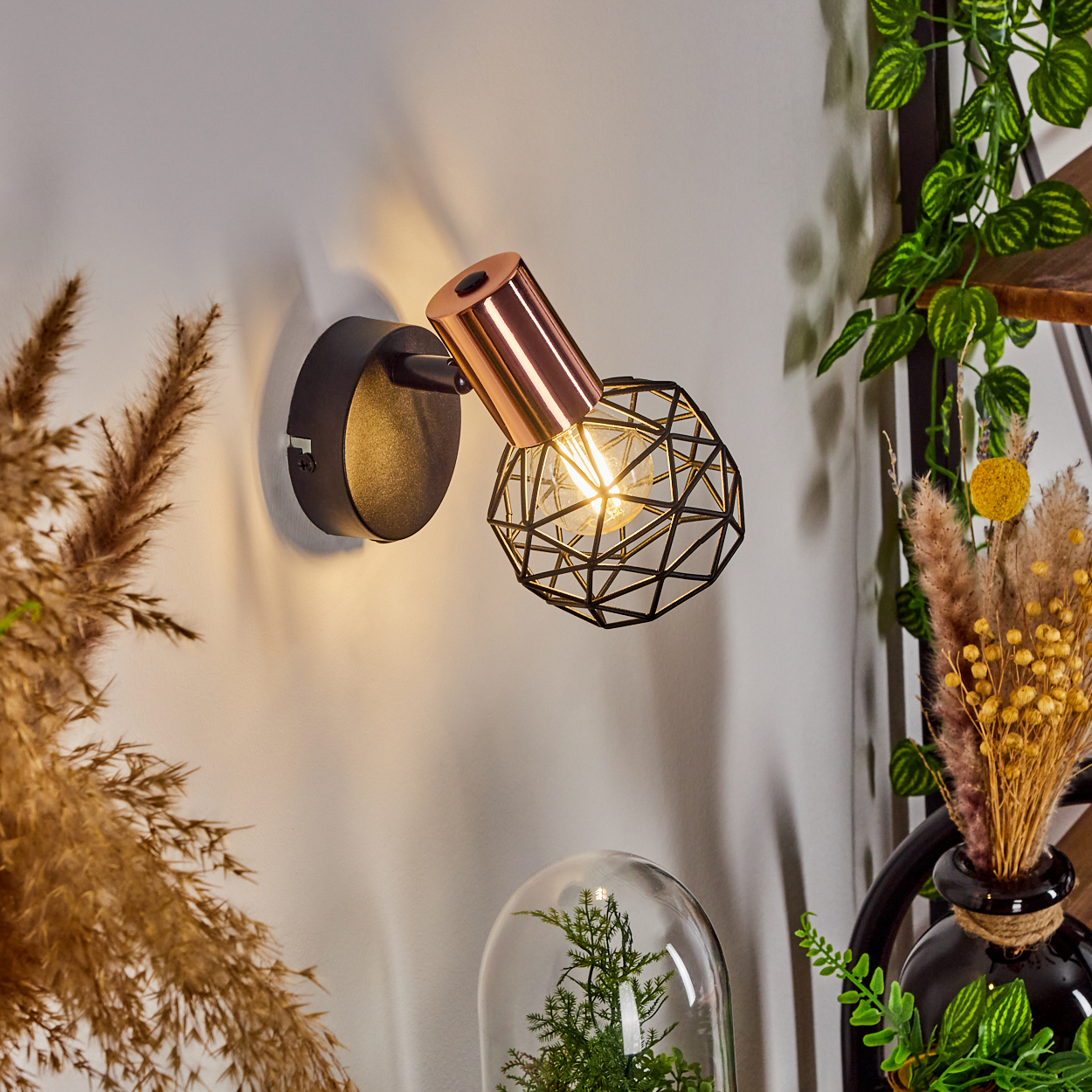 Eine Wandlampe mit schwarzem Rahmen und kupferfarbener Lichtquelle. Der Hintergrund ist eine weiße Wand.
