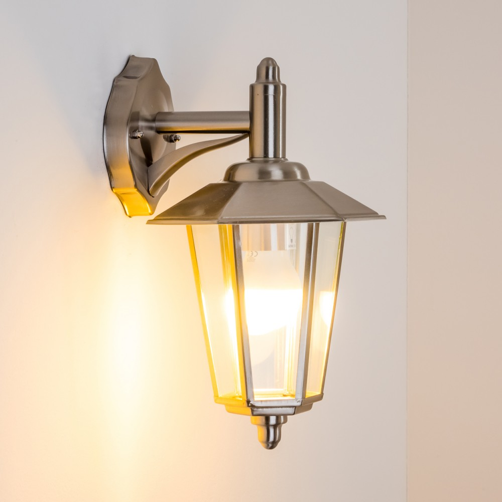 Eine silberne Außenwandleuchte mit Glasgehäuse. Sie ist an einer weißen Wand montiert.