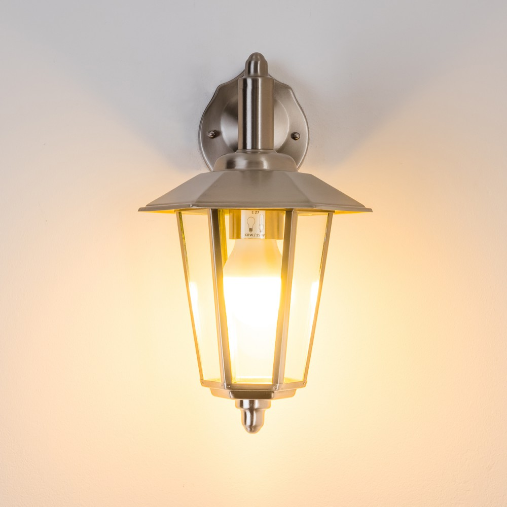 Eine an der Wand montierte Außenlampe mit Glasgehäuse. Die Glühbirne leuchtet, die Wand ist weiß.