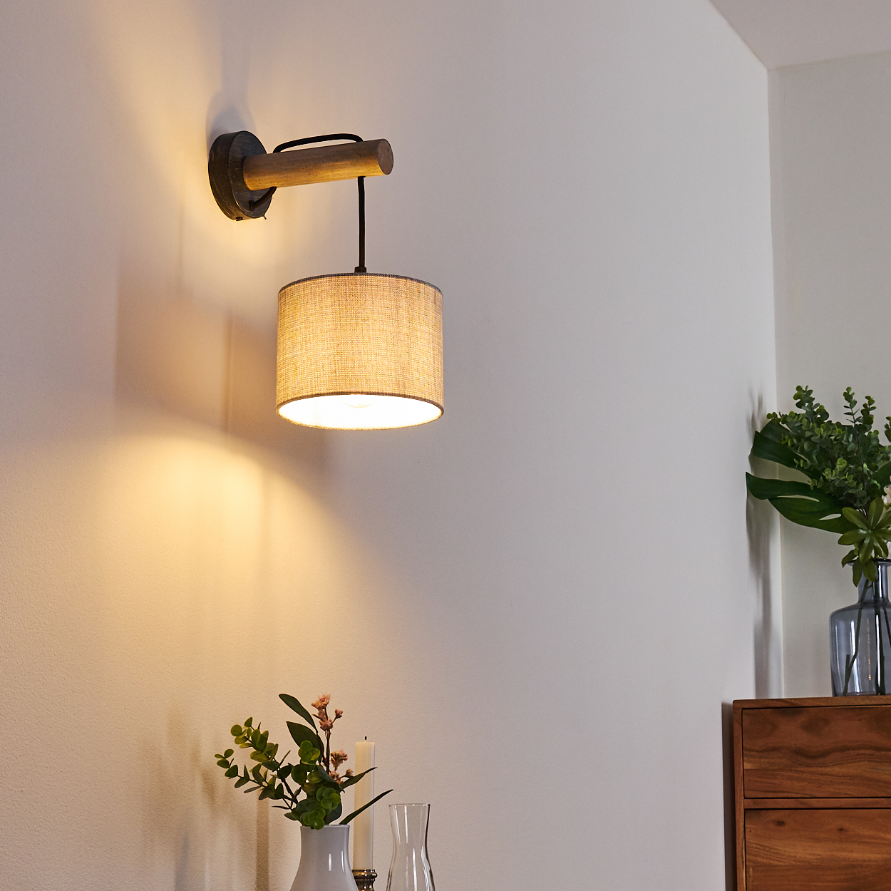 Eine Wandleuchte mit beigefarbenem Lampenschirm, der an einem Holzbogen hängt. Die Wand ist weiß.