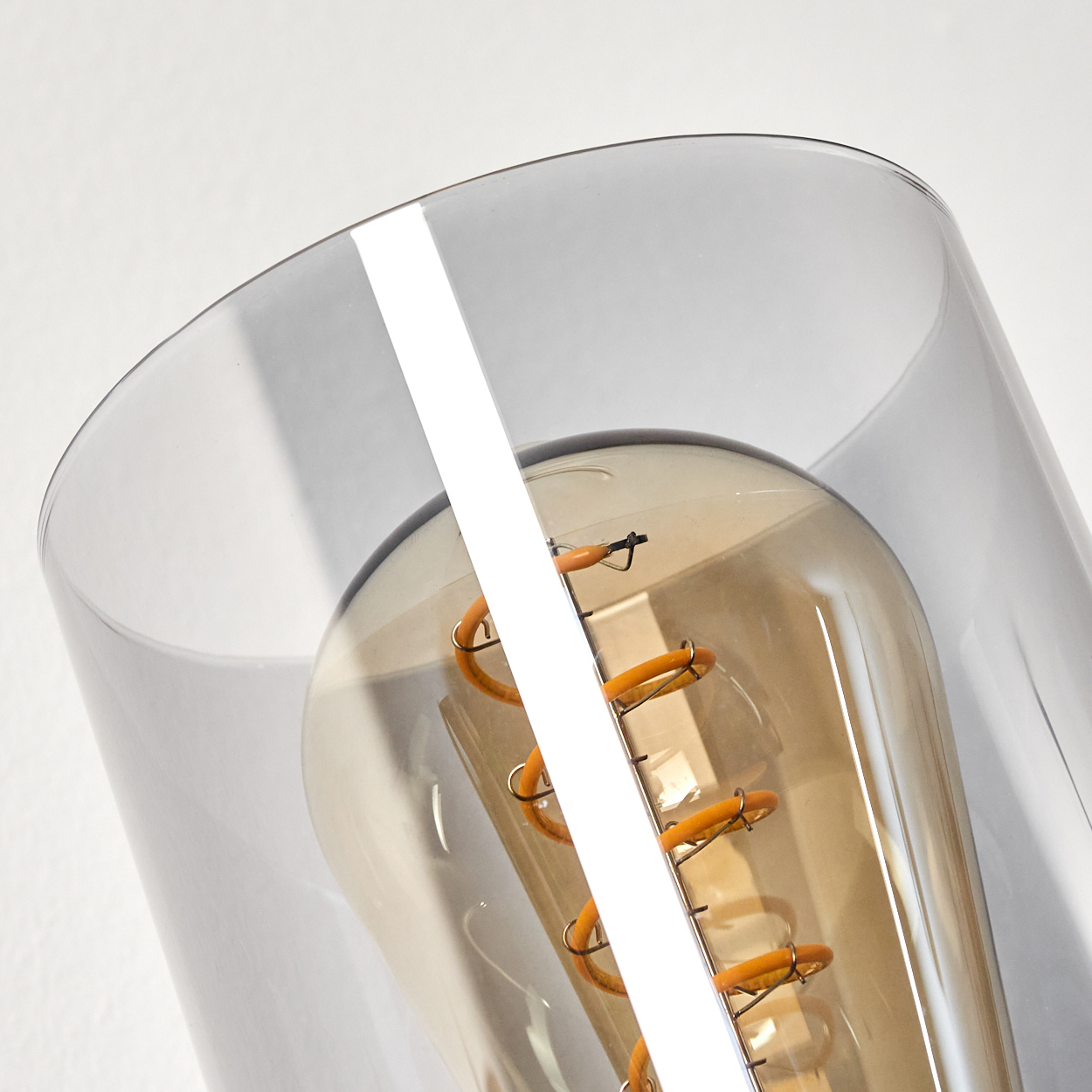 Nahaufnahme einer Glühbirne in einem transparenten, zylindrischen Glasschirm. Weißer Wandhintergrund.