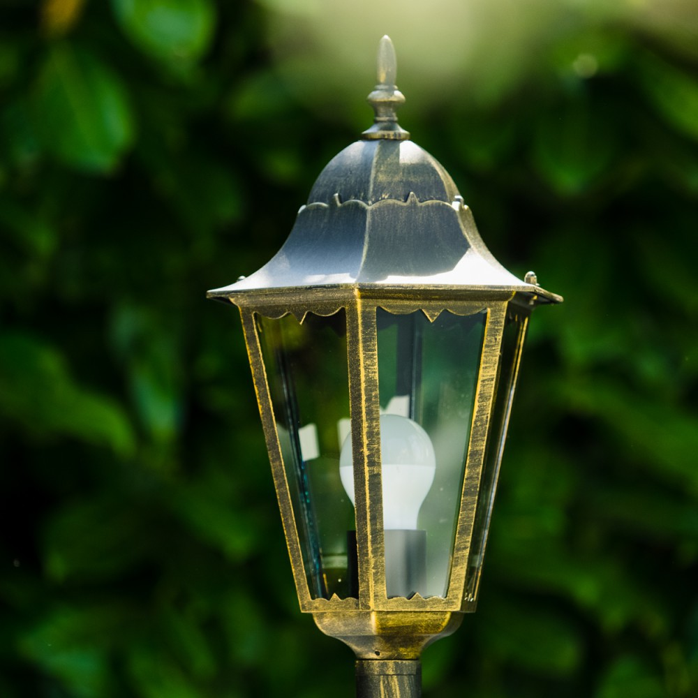 Eine Außenlaterne mit goldenem Oberteil und hellem Licht im Inneren. Grünes Laub im Hintergrund.