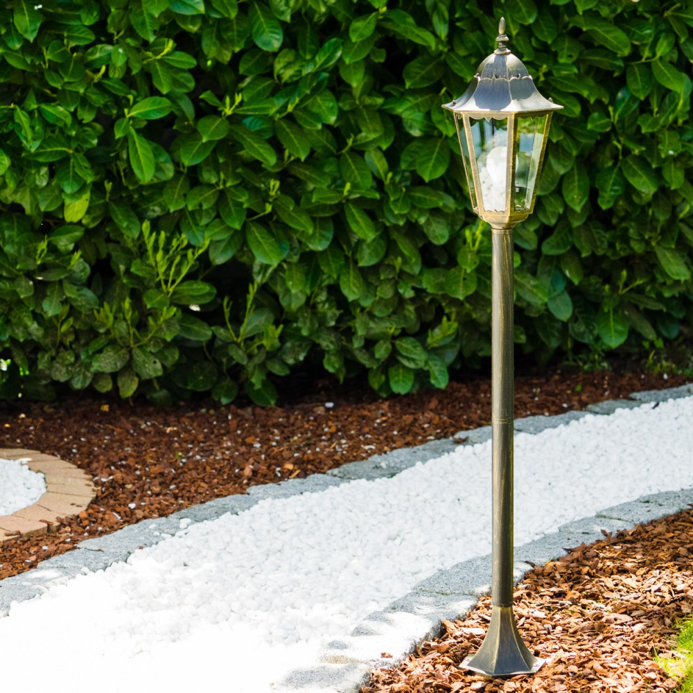 Eine Außenlaterne mit brennender Glühbirne. Die Lampe ist bronze- und grünfarben und steht auf einem Weg.