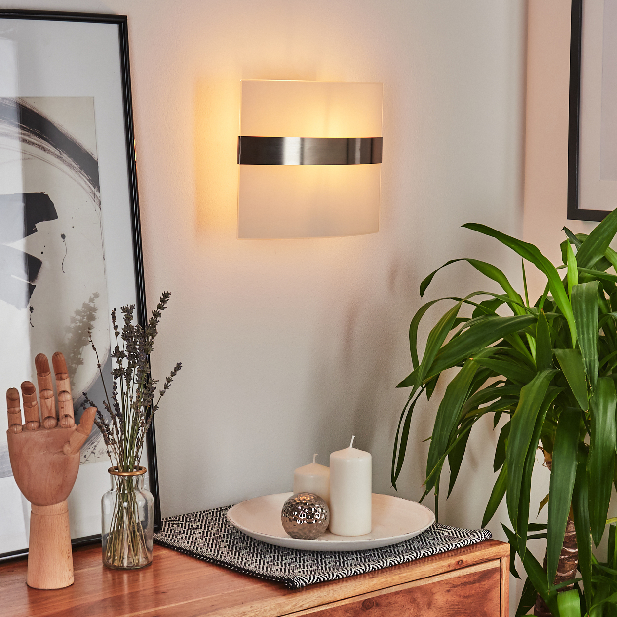 Eine Wandlampe mit Milchglasschirm und silbernen Akzenten, in der Nähe eines Holztisches mit Deko.
