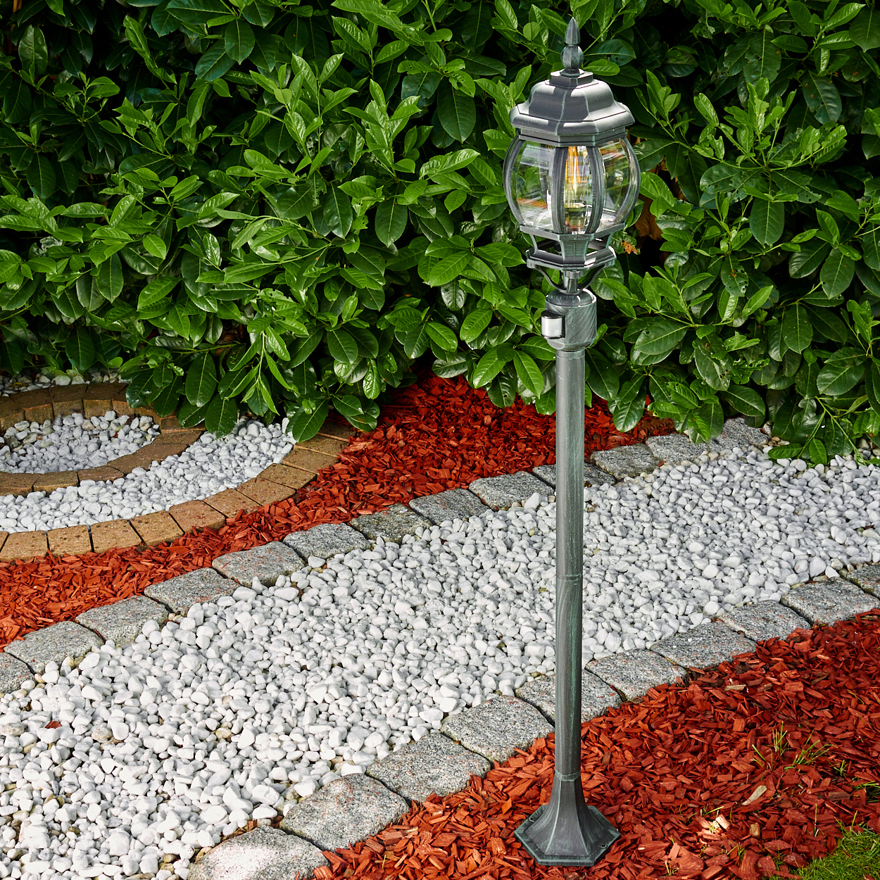 Laternenpfahl im Garten. Er hat einen klaren Glasschirm und ist von Pflanzen umgeben.