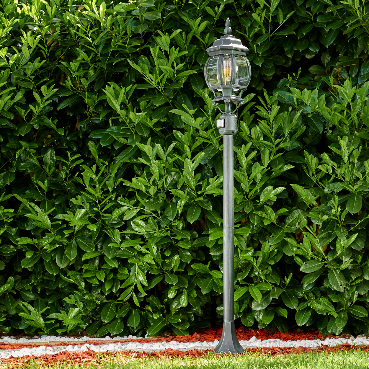 Eine graue Außenlampe steht vor einer grünen Hecke und verleiht der Gartenanlage Licht und Charme.