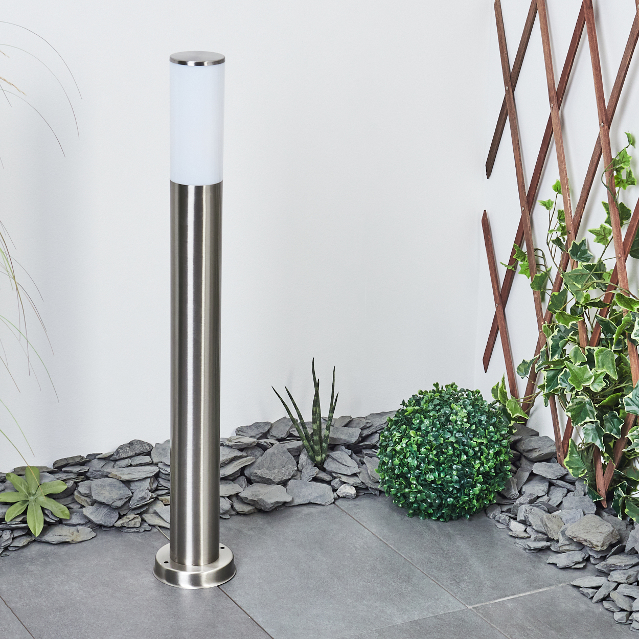 Außenleuchte in einem Garten. Sie hat eine weiße Lichtsäule und einen silbernen Sockel. Hintergrund: Stein und Pflanzen.
